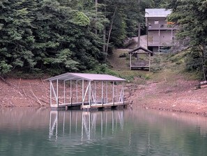 Un muelle cubierto se adentra en un lago tranquilo, rodeado de un bosque frondoso y un pequeño pabellón.