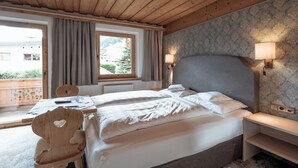 A bedroom with a wooden ceiling, a bed with white linens, a wooden nightstand, a desk with a chair, and a window with curtains. - Gästehaus Wildschütz (Tux)