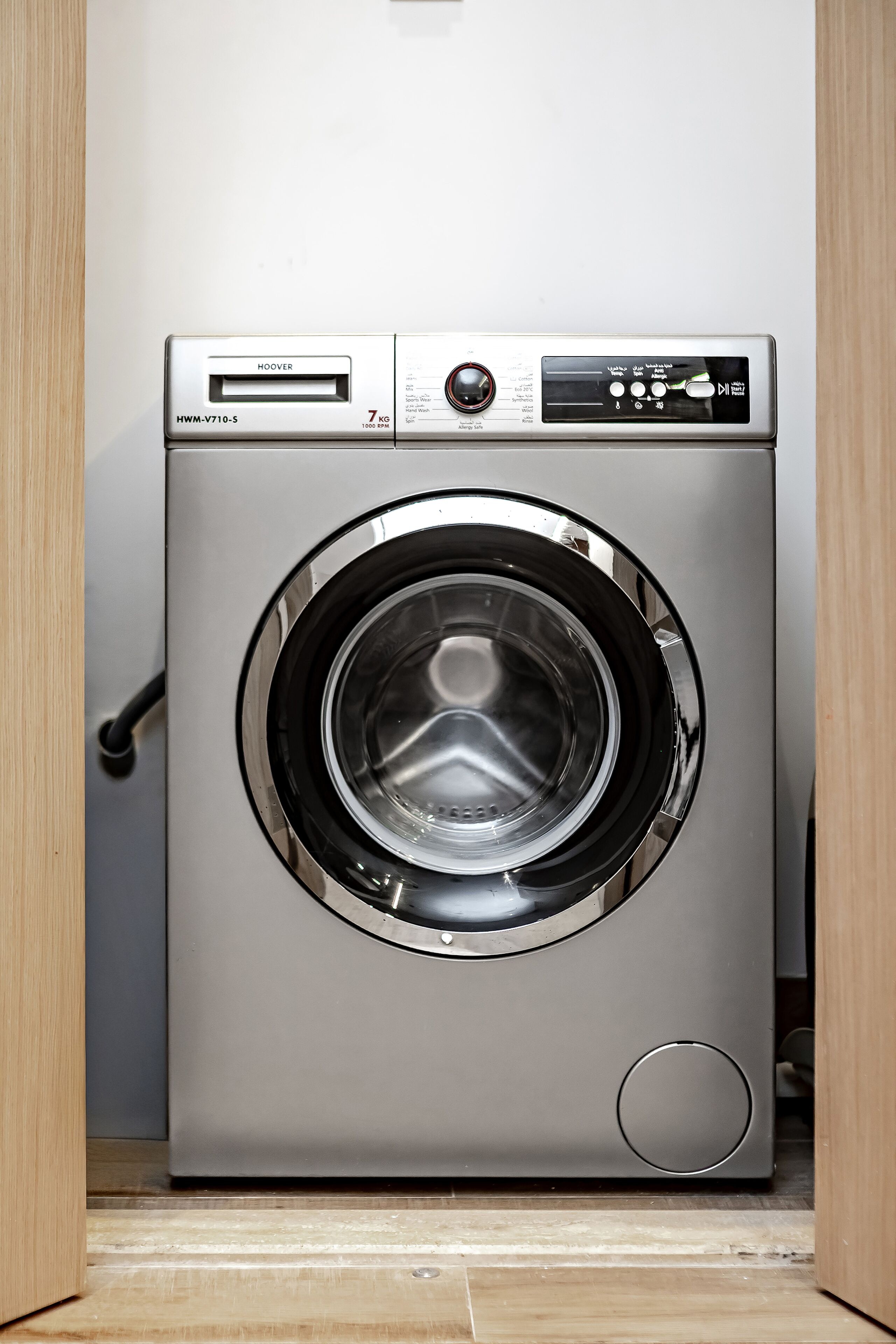 A front view of a compact, stainless steel front-loading washing machine with a digital control panel, located in a wooden-framed laundry room.