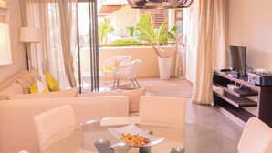 Een moderne woonkamer met een glazen tafel, witte stoelen, een bank met gele kussens, een televisie en een balkon met planten.