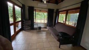 A spacious room with a tiled floor, a ceiling fan, a sofa, and large windows offering a view of greenery outside.