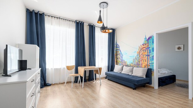 Uma sala de estar moderna com piso de madeira, um sofá azul, uma mesa de jantar com duas cadeiras, uma TV em um móvel branco e um mural colorido com vista para a cidade.