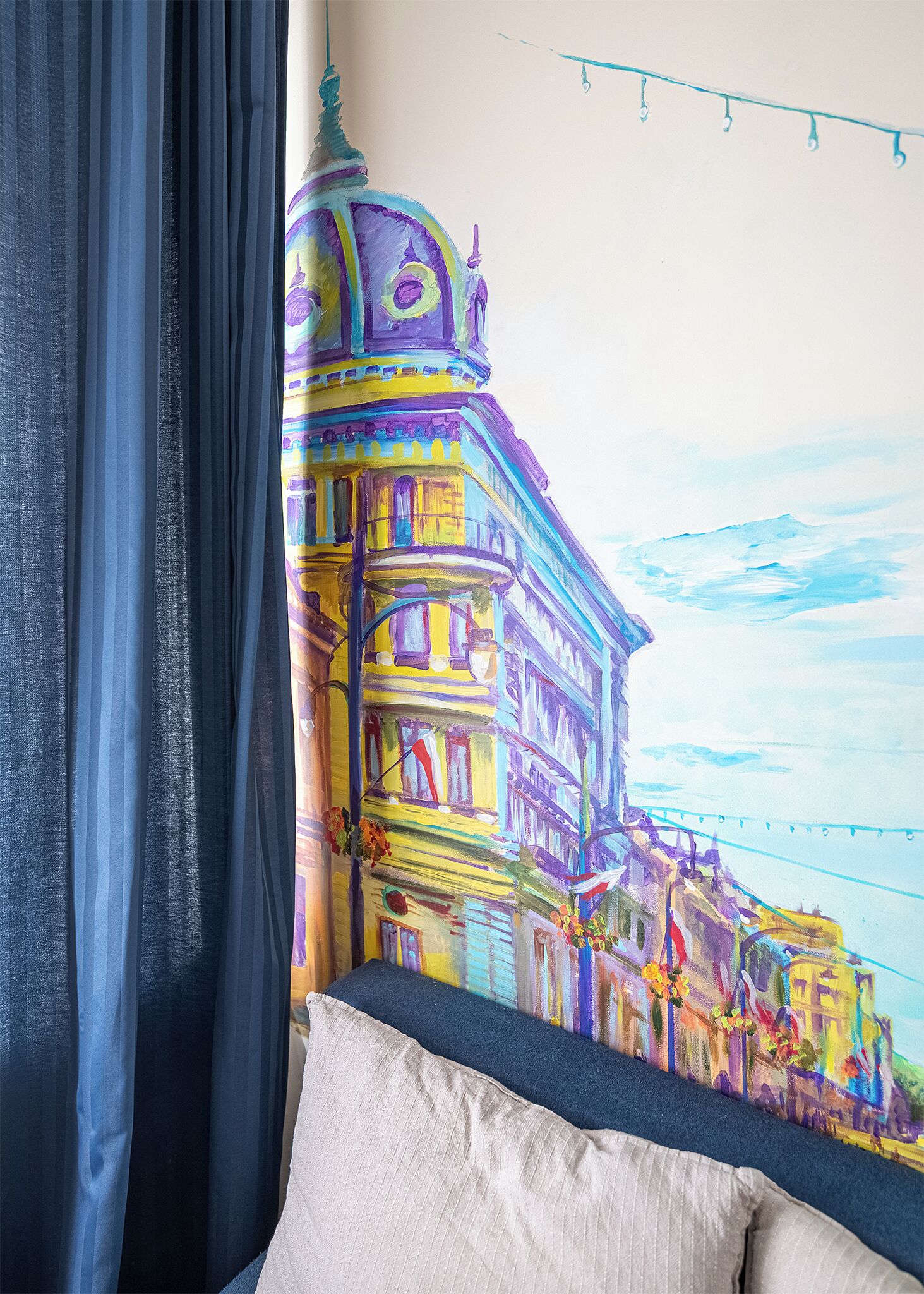Una habitación con un mural colorido de un edificio, una cama con un almohadón blanco y una guirnalda de luces.