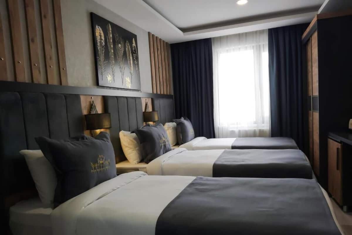 Ein Hotelzimmer mit zwei Betten, einem großen Fenster mit Vorhängen, einem hölzernen Kopfteil und einem an der Wand befestigten Kunstwerk.