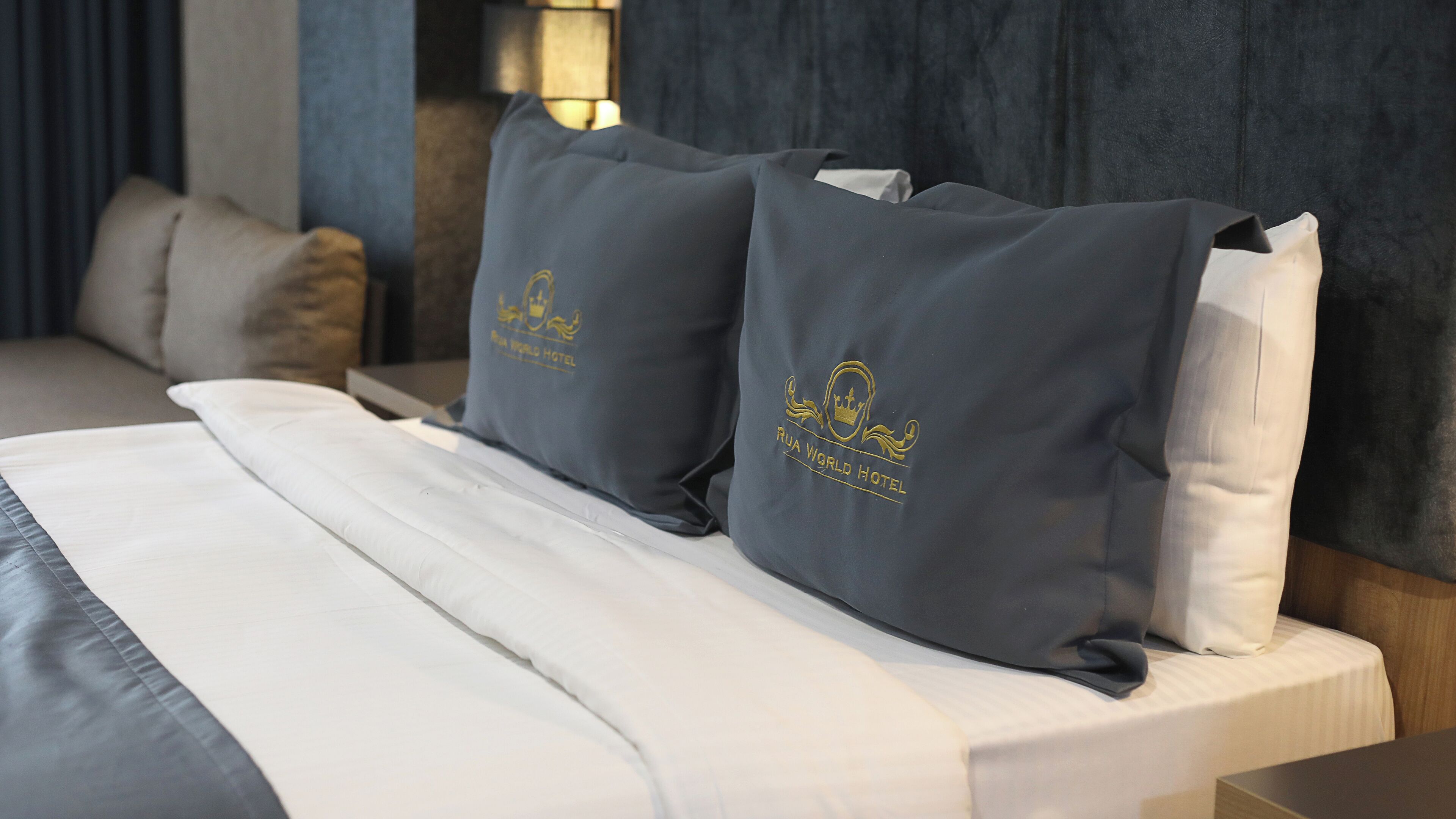 A bed with white linens and two pillows with the RUA WORLD HOTEL logo.