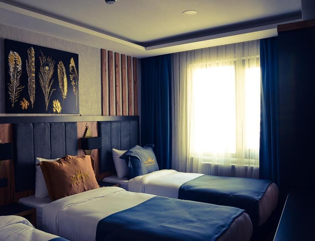 A hotel room with two beds, a headboard with vertical slats, a large window with curtains, and a wall-mounted artwork of feathers.
