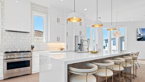 Una cucina moderna con mobili bianchi, una grande isola con sgabelli da bar, elettrodomestici in acciaio inossidabile e illuminazione a sospensione.