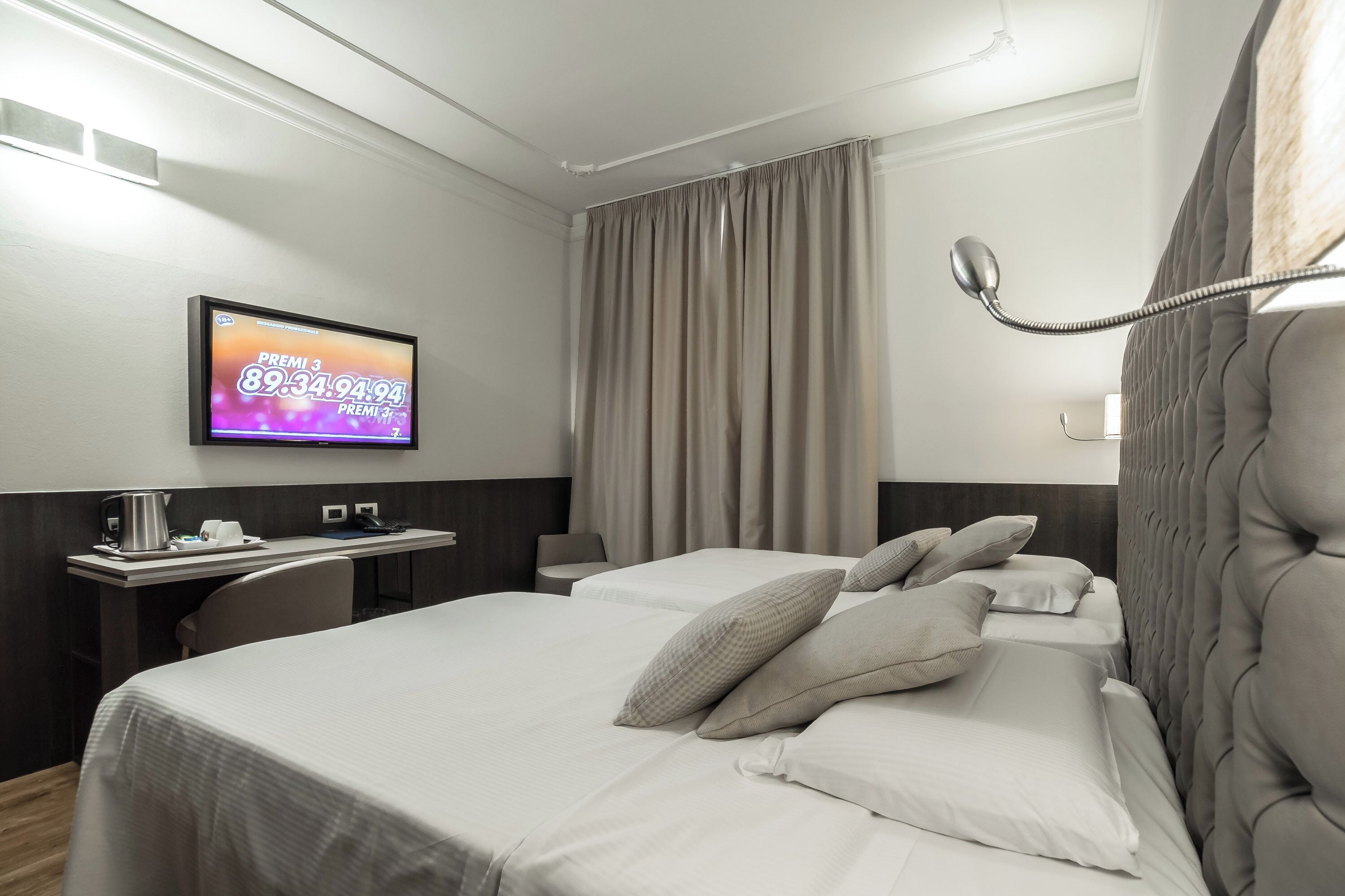 Ein Hotelzimmer mit einem Bett, einem Schreibtisch mit Kaffeemaschine, einem Fernseher, der ein Sportereignis zeigt, und einem großen Fenster mit Vorhängen.