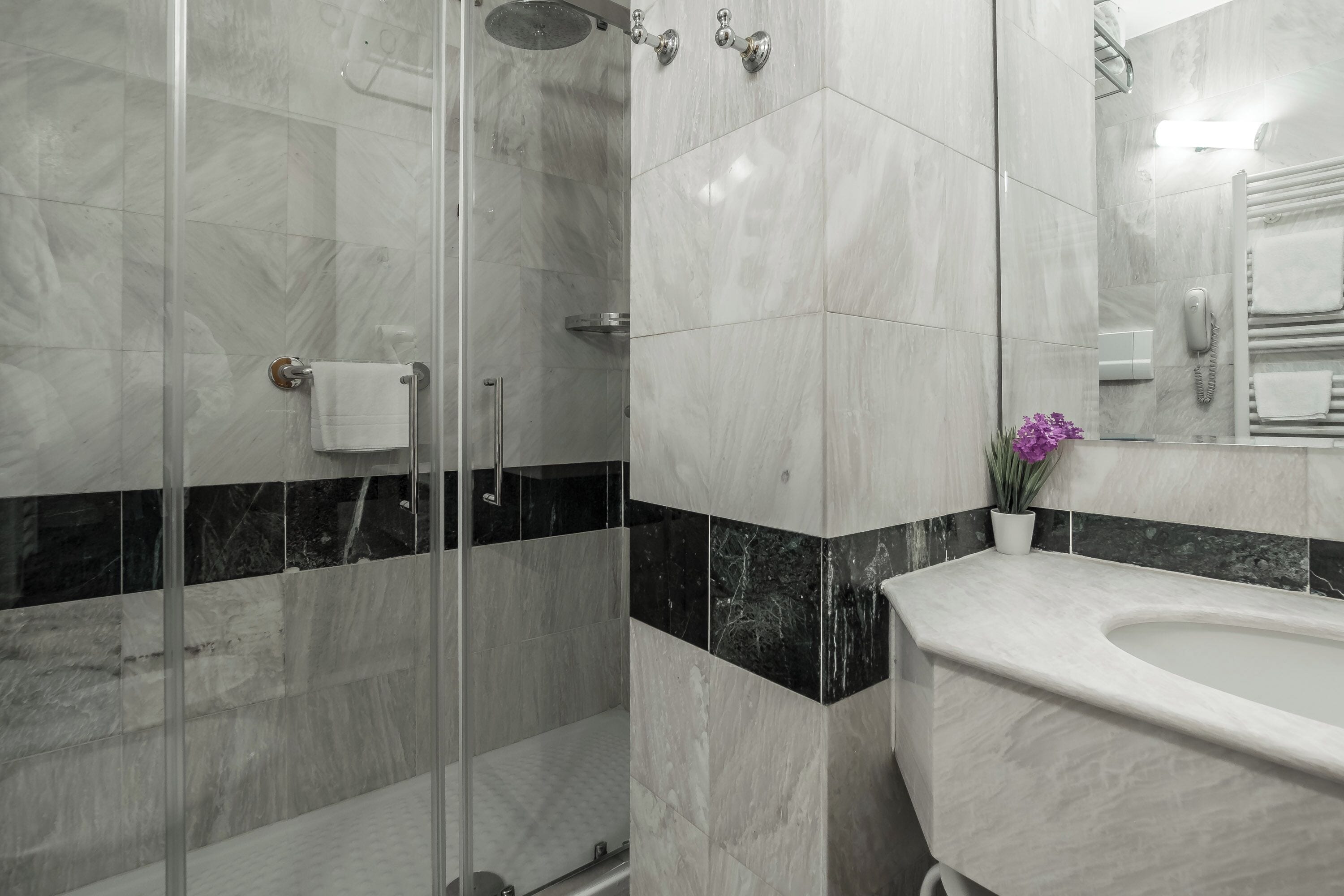 A modern bathroom with a glass shower enclosure, a marble-tiled shower area, a white sink, and a mirror with a towel rack.