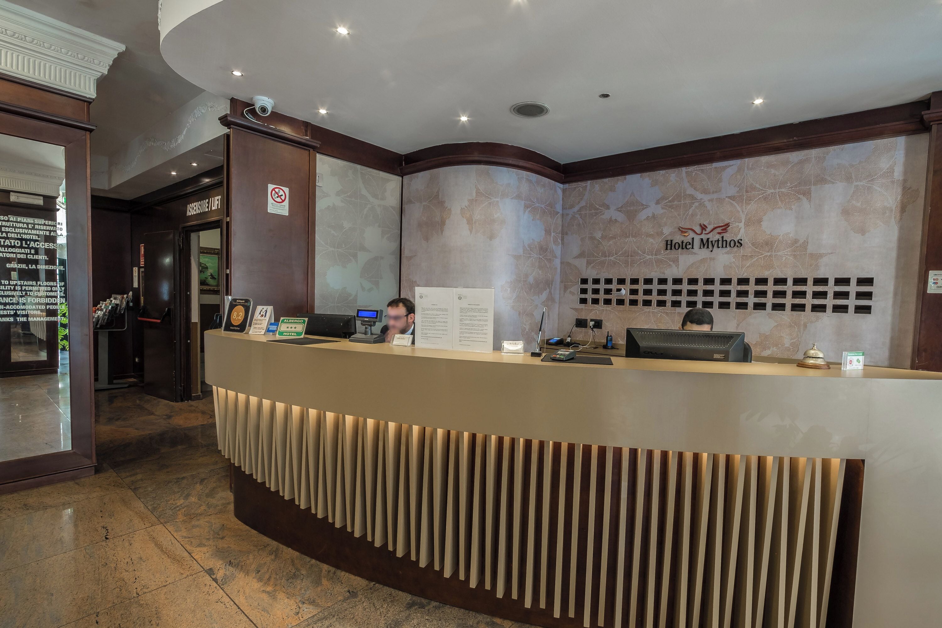 A hotel reception desk with a modern design, a sign indicating no smoking, and a logo for Hotel Mythos.