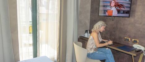 A hotel room with a bed, a desk with a laptop, a TV on the wall, and a window with sheer curtains.