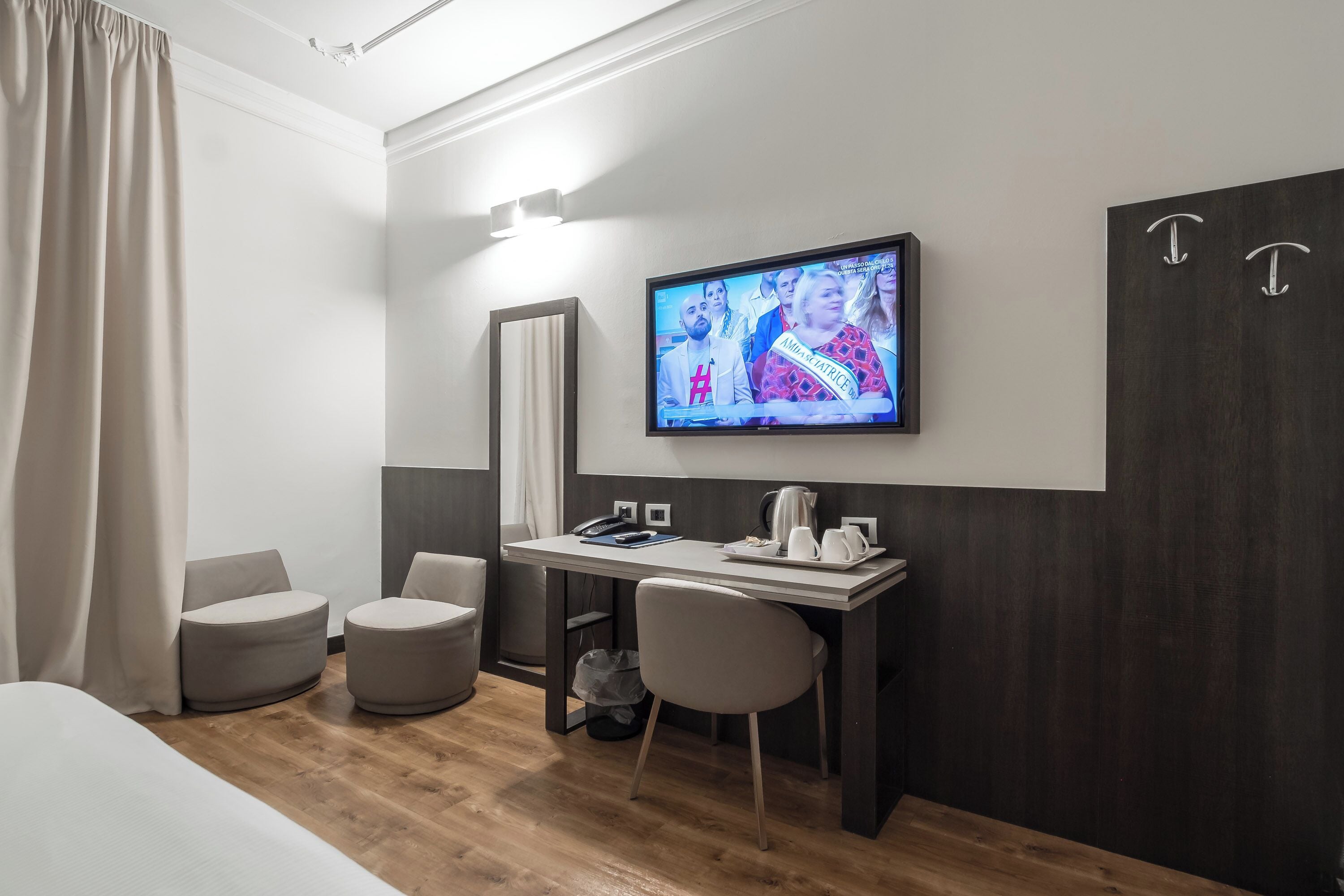 A modern hotel room with a wooden floor, a desk with a chair, a television, a large mirror, and a bed.