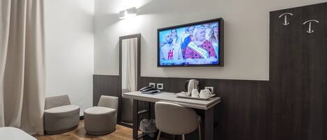 A modern hotel room with a wooden floor, a desk with a chair, a television, a large mirror, and a bed.