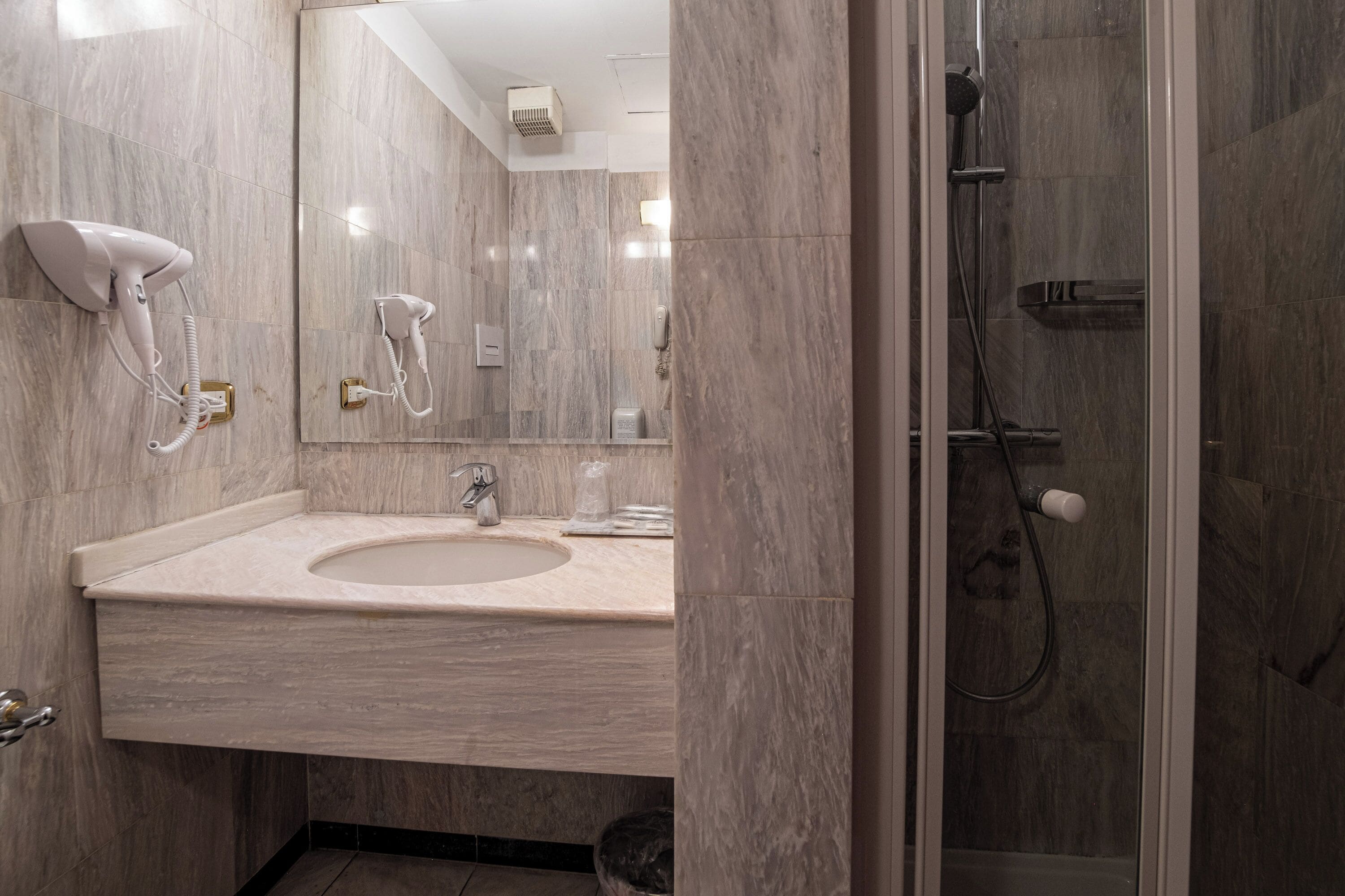 A modern bathroom with a marble sink, a hairdryer, a mirror, and a shower enclosure.