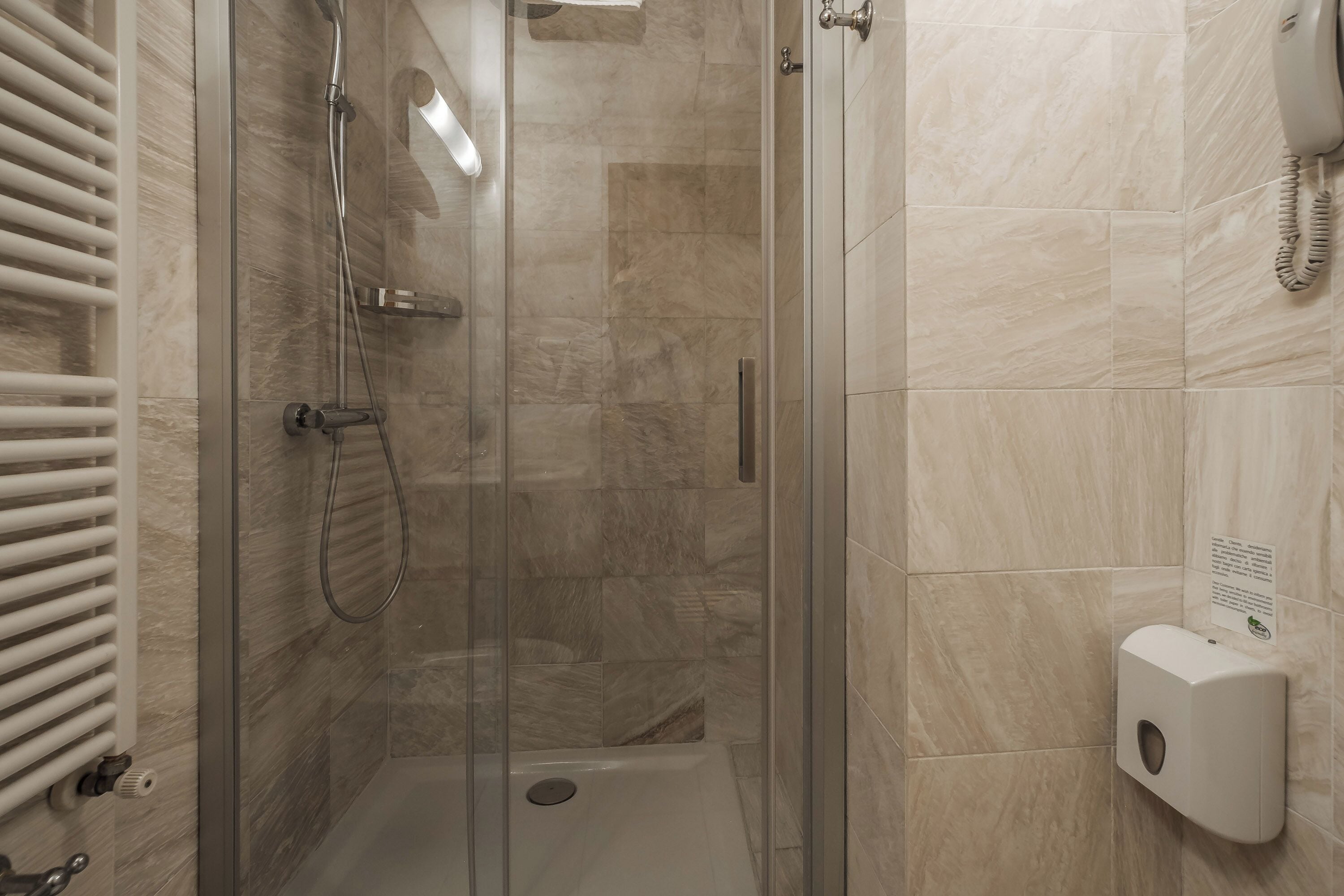 A glass-enclosed shower stall with a handheld showerhead, a wall-mounted toilet paper dispenser, and a towel rack with white towels.