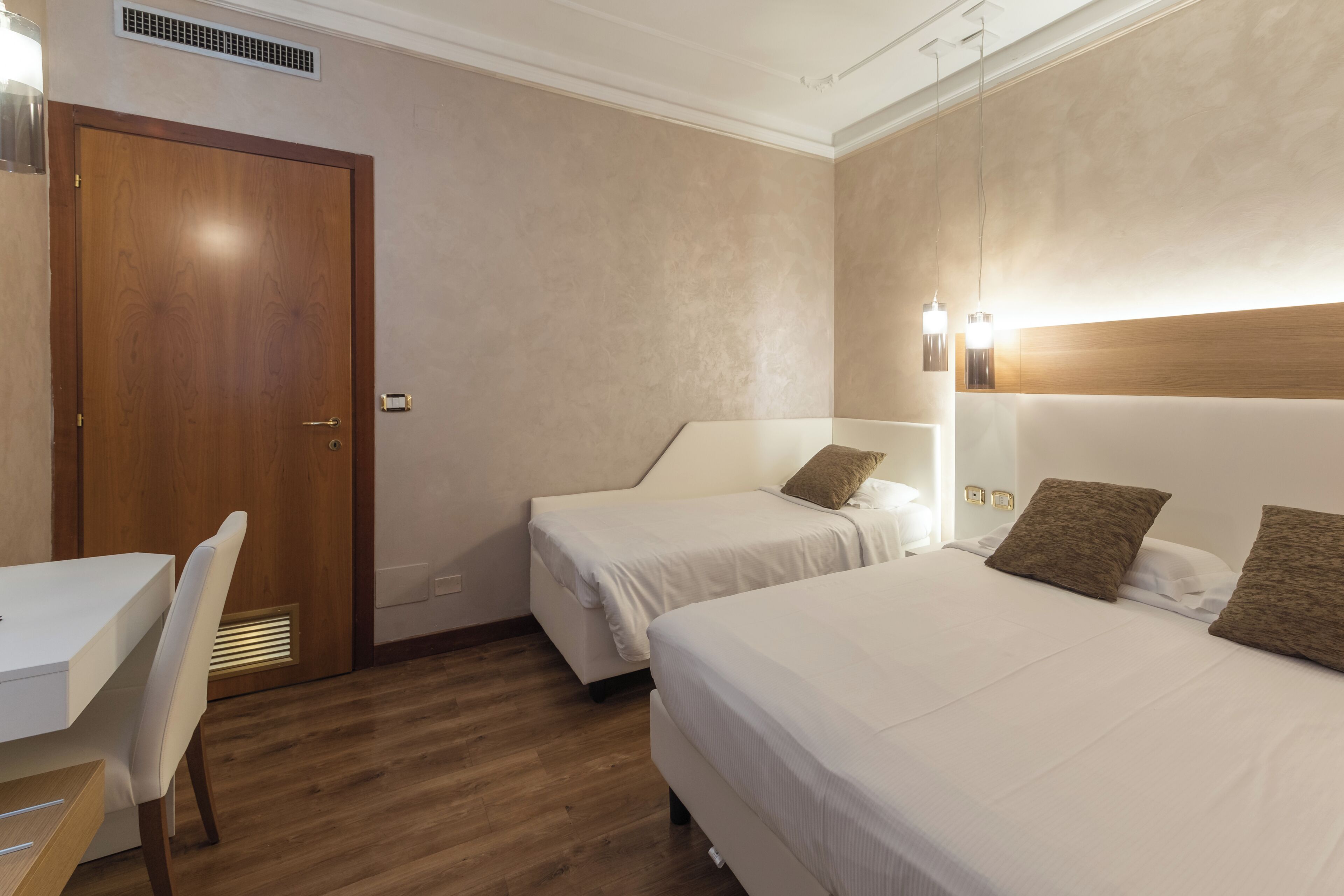 A hotel room with two single beds, a wooden door, a white desk, and a chair.