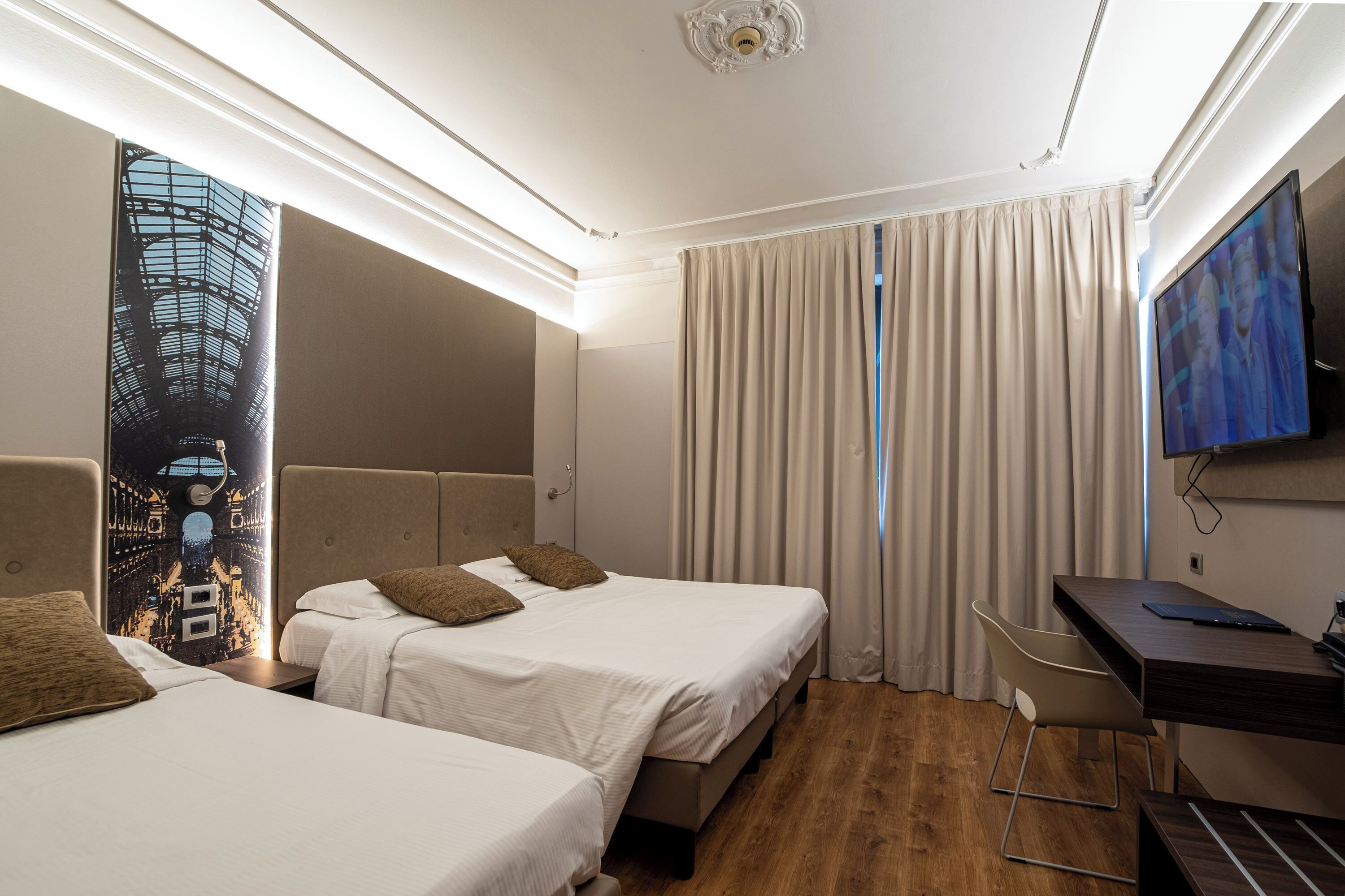 A hotel room with two beds, a desk, a chair, a TV, and a large window with curtains.