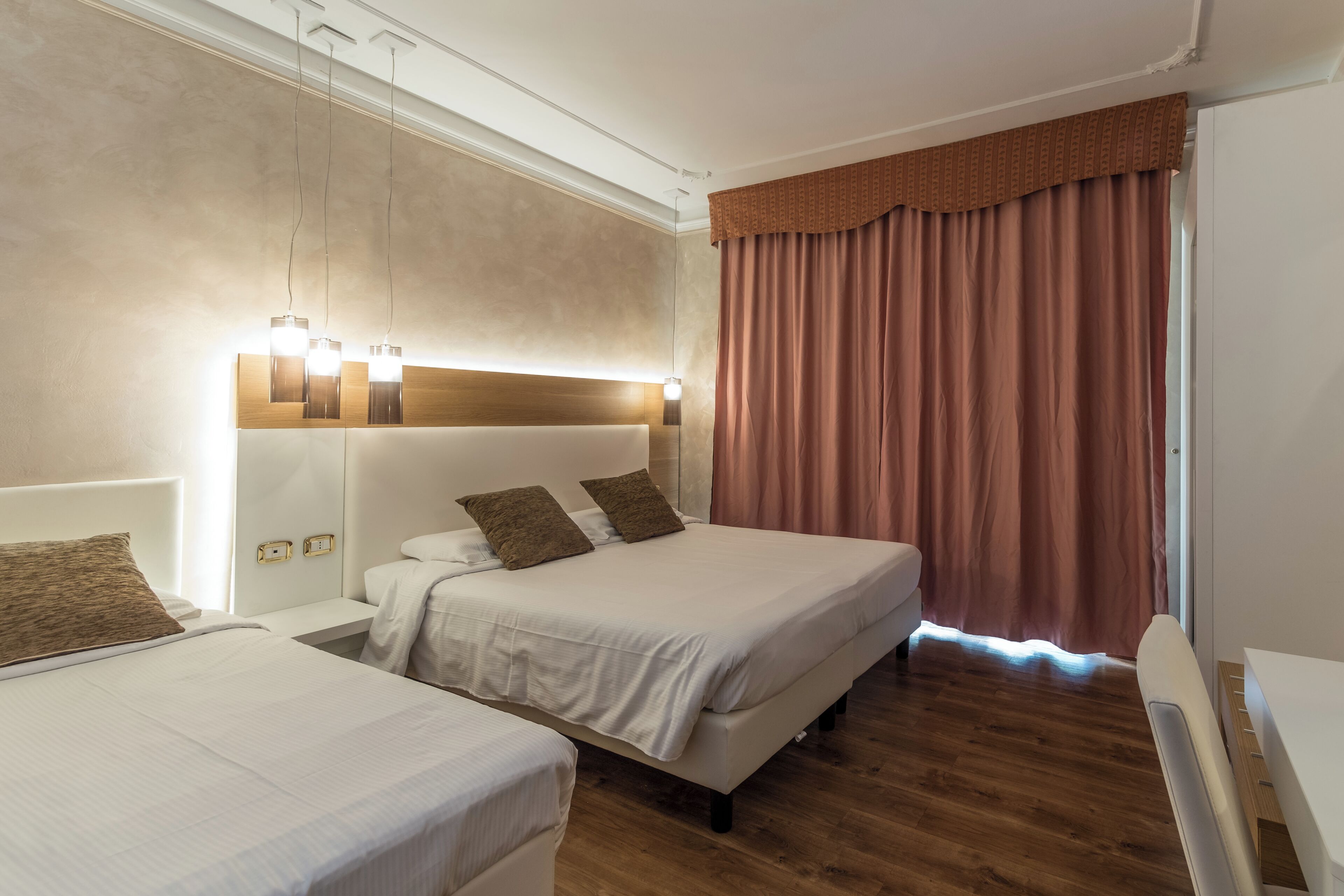 Ein Hotelzimmer mit zwei Betten, Holzboden, einem großen Fenster mit Vorhängen und einem modernen Kopfteil mit integrierter Beleuchtung.