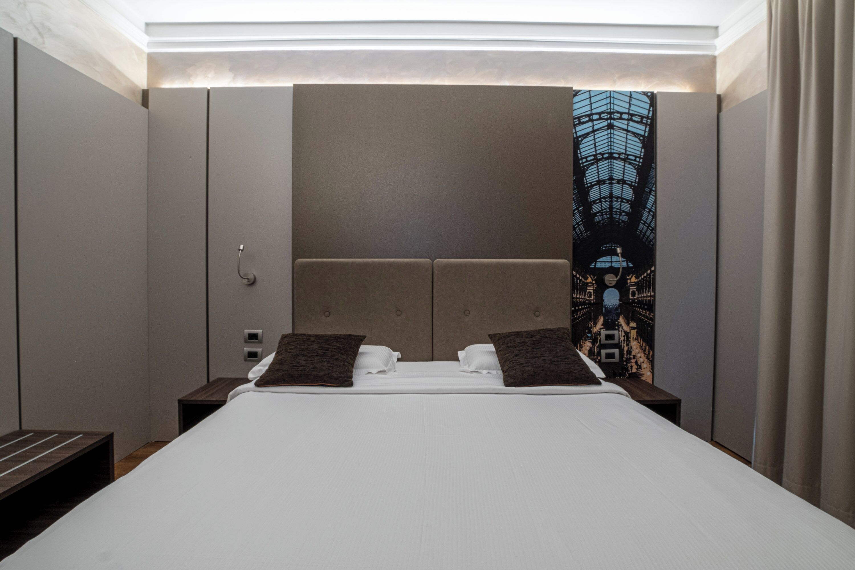 Une chambre d’hôtel moderne avec un grand lit, une tête de lit ornée d’une fresque représentant un paysage urbain, une table de chevet et une petite table de nuit.