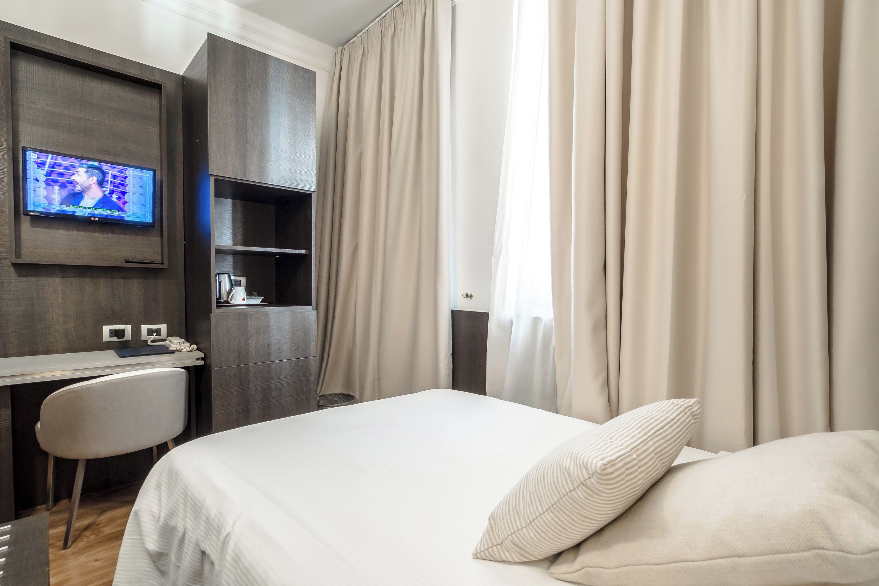 A hotel room with a bed, a desk, a television mounted on the wall, and a window with curtains.