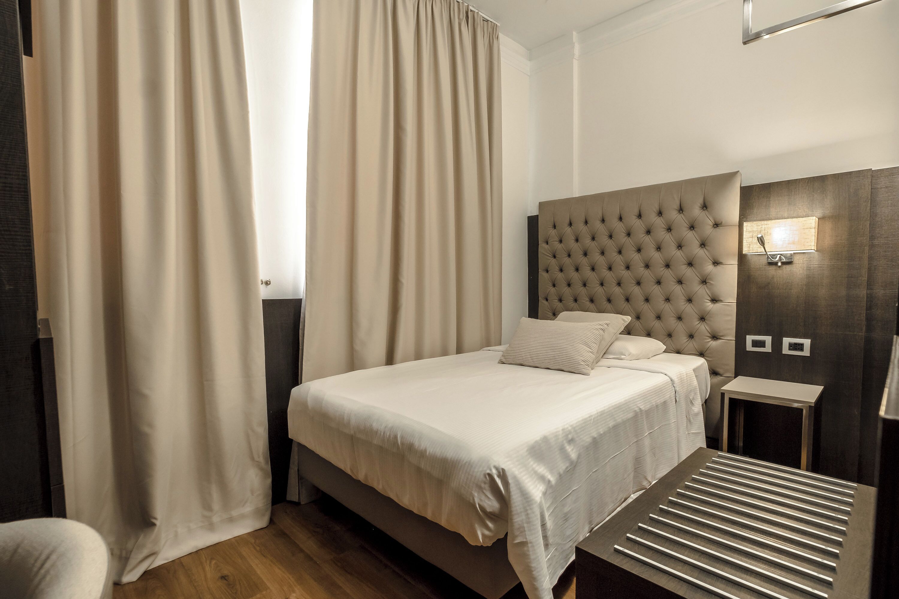 A neatly arranged hotel room with a large bed, a bedside table, a wall-mounted light, and a window with curtains.