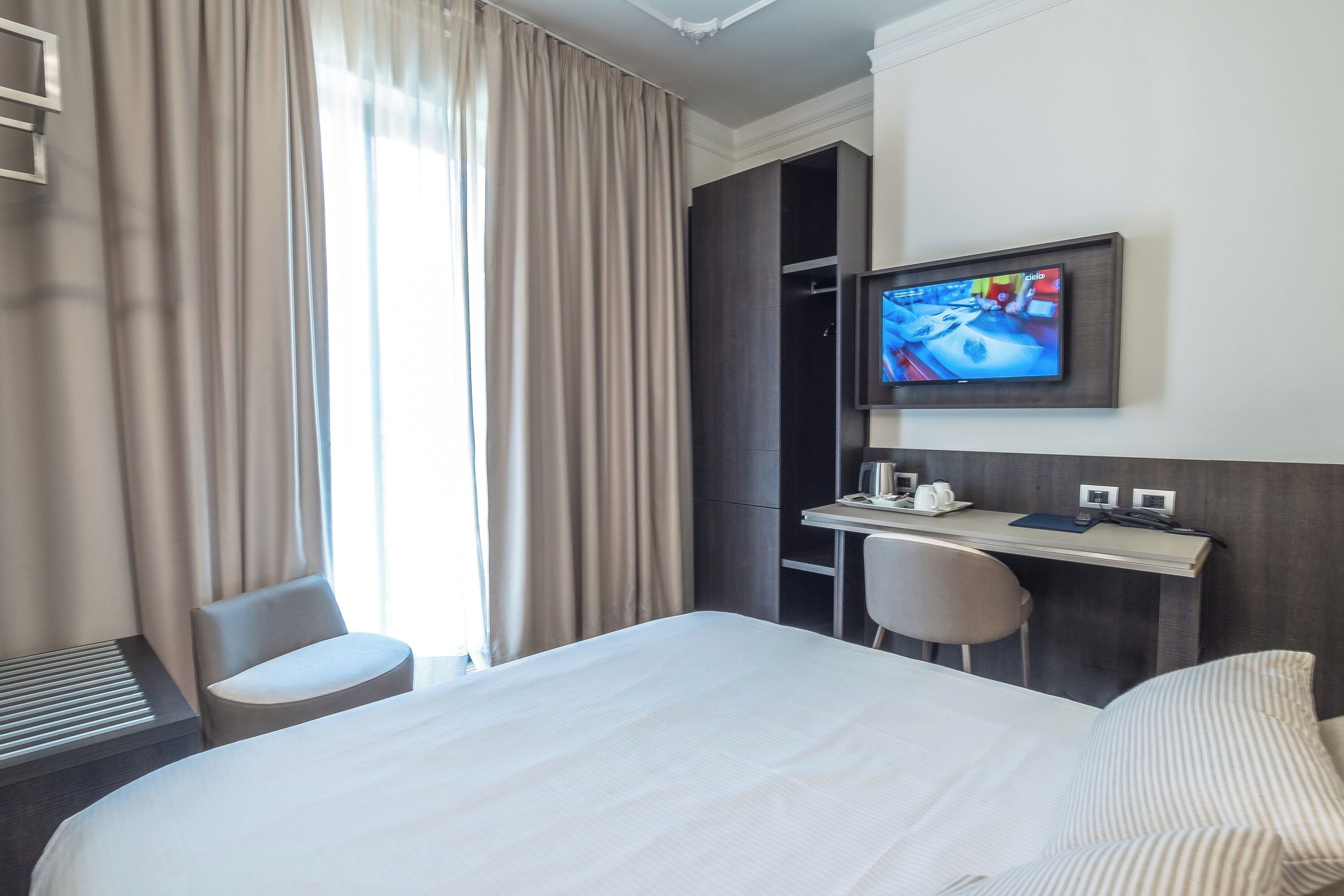 Une chambre d’hôtel avec un grand lit, un bureau avec une chaise, un téléviseur fixé au mur et une fenêtre avec des rideaux.