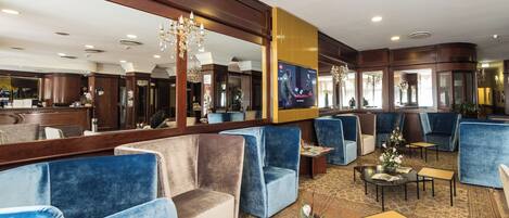 Un vestíbulo de hotel con asientos en tonos azul y beige, un mostrador de recepción y una gran pantalla de televisión.