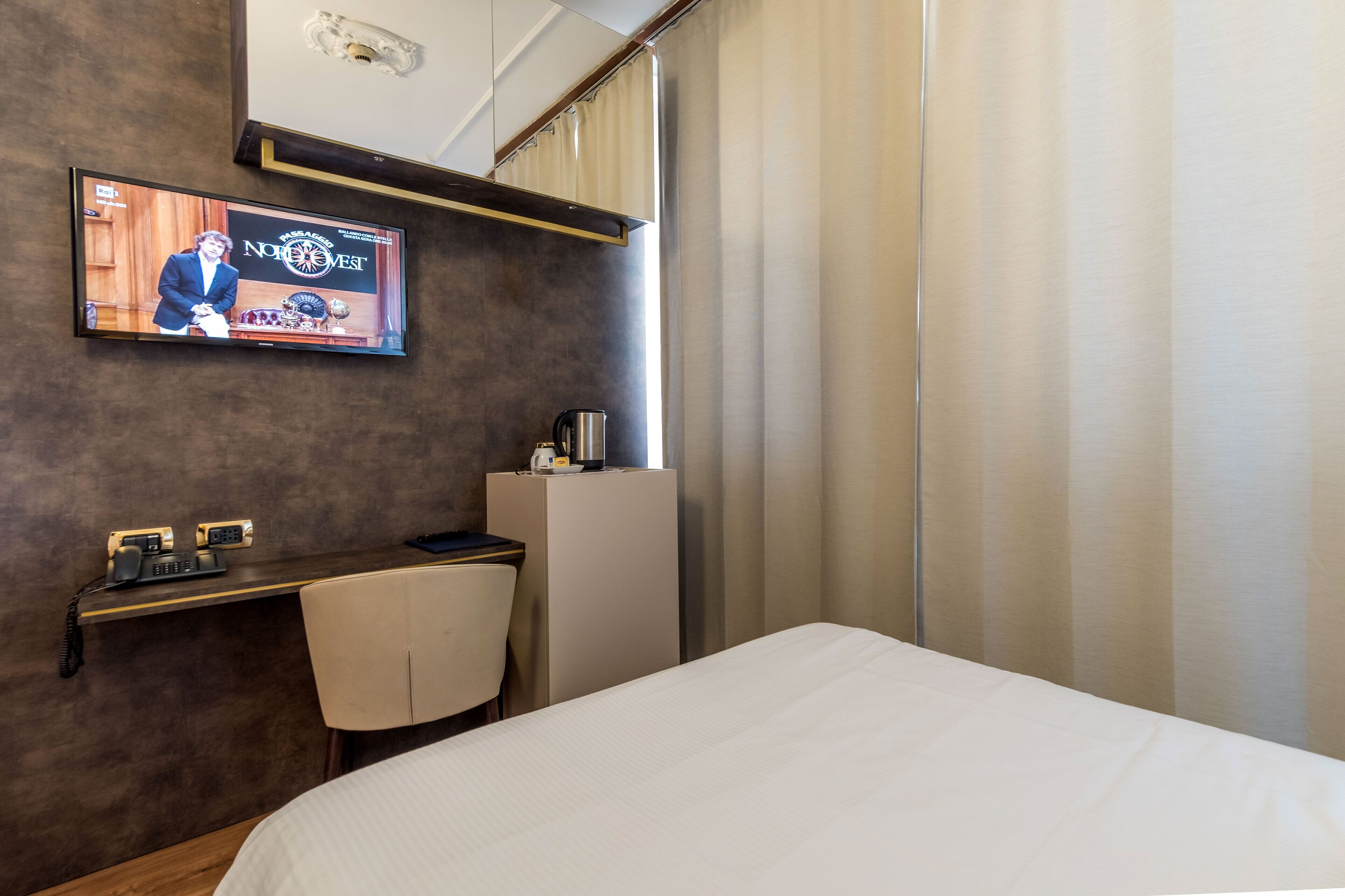 A hotel room with a bed, a desk with a chair, a television mounted on the wall, and a window with curtains.