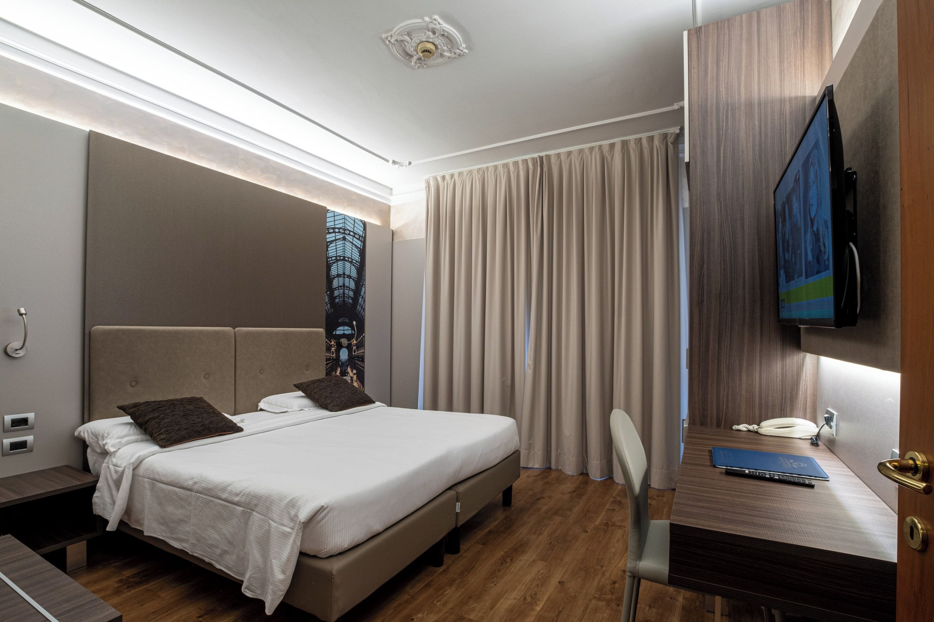 A modern hotel room with a large bed, a desk with a laptop, a television, and a view of a building through the window.