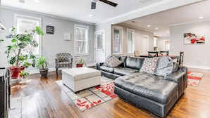 A spacious living room with a large black leather sofa, a white ottoman, and a ceiling fan. There are potted plants and a dining area in the background. - 4 Mi to Dtwn: Relaxing Escape w/ Yard in Atlanta (Atlanta)