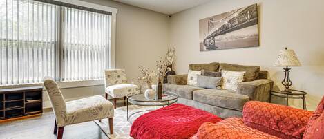 A living room with a sofa, armchair, and coffee table. There is a window with blinds, a painting on the wall, and a lamp on a side table.