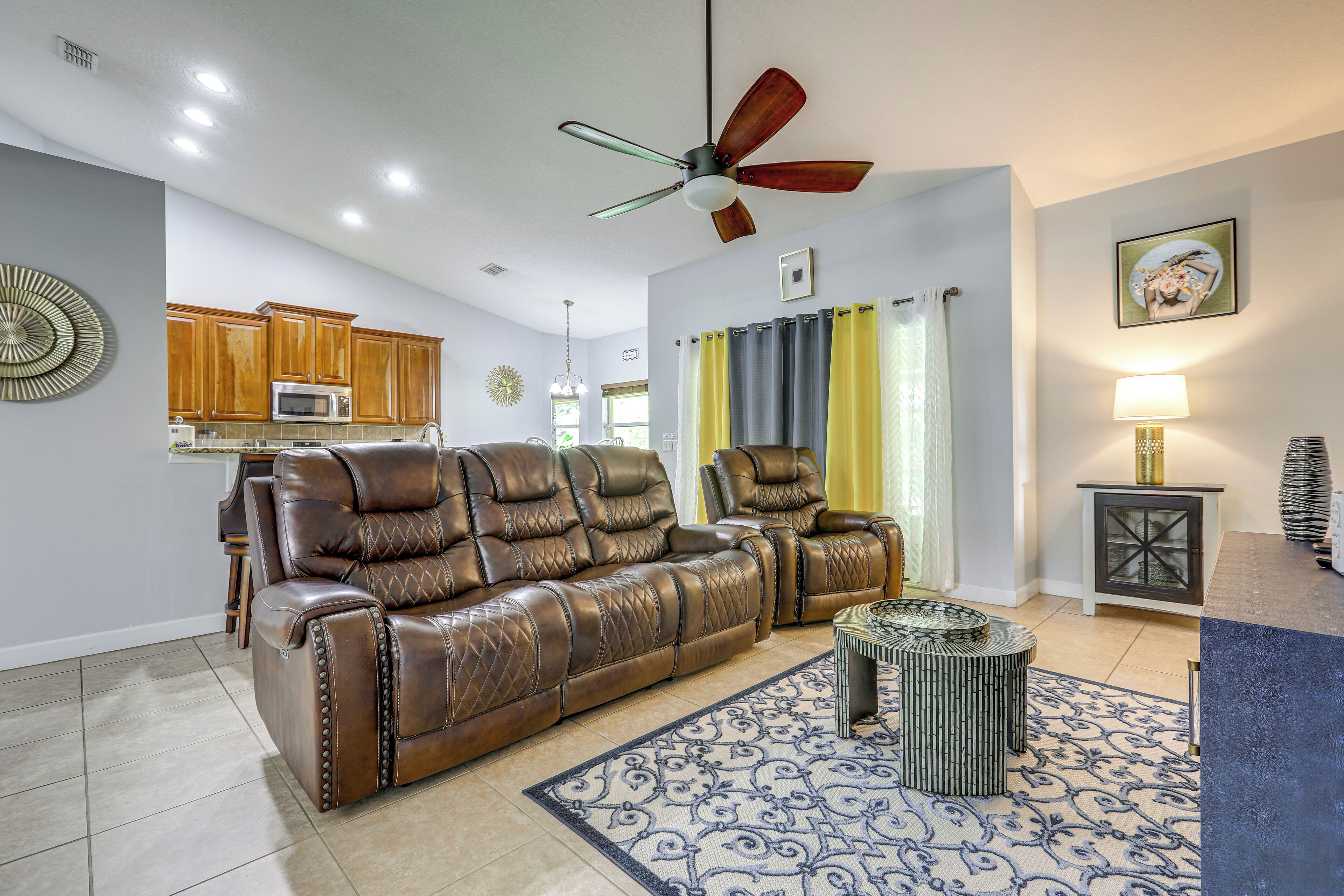 A living room with a brown leather sofa, a ceiling fan, a patterned rug, and a kitchen area in the background.