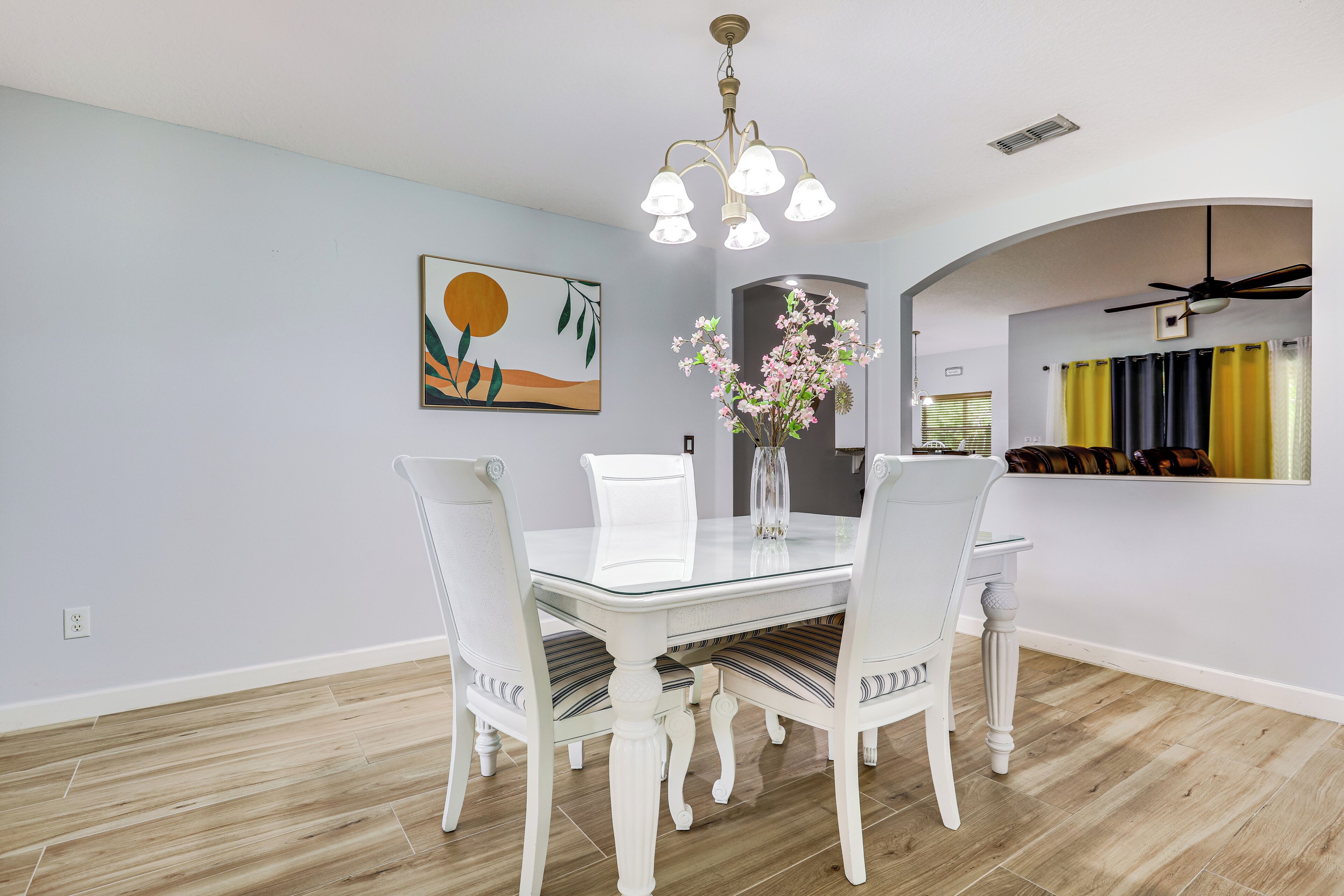 A dining area with a glass table and white chairs, a chandelier, and a painting on the wall.