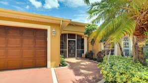 A residential house with a garage, a covered entryway, and a well-maintained garden with palm trees and shrubs. - Family Home in Port St Lucie w/ Furnished Pergola! (Port Saint Lucie)