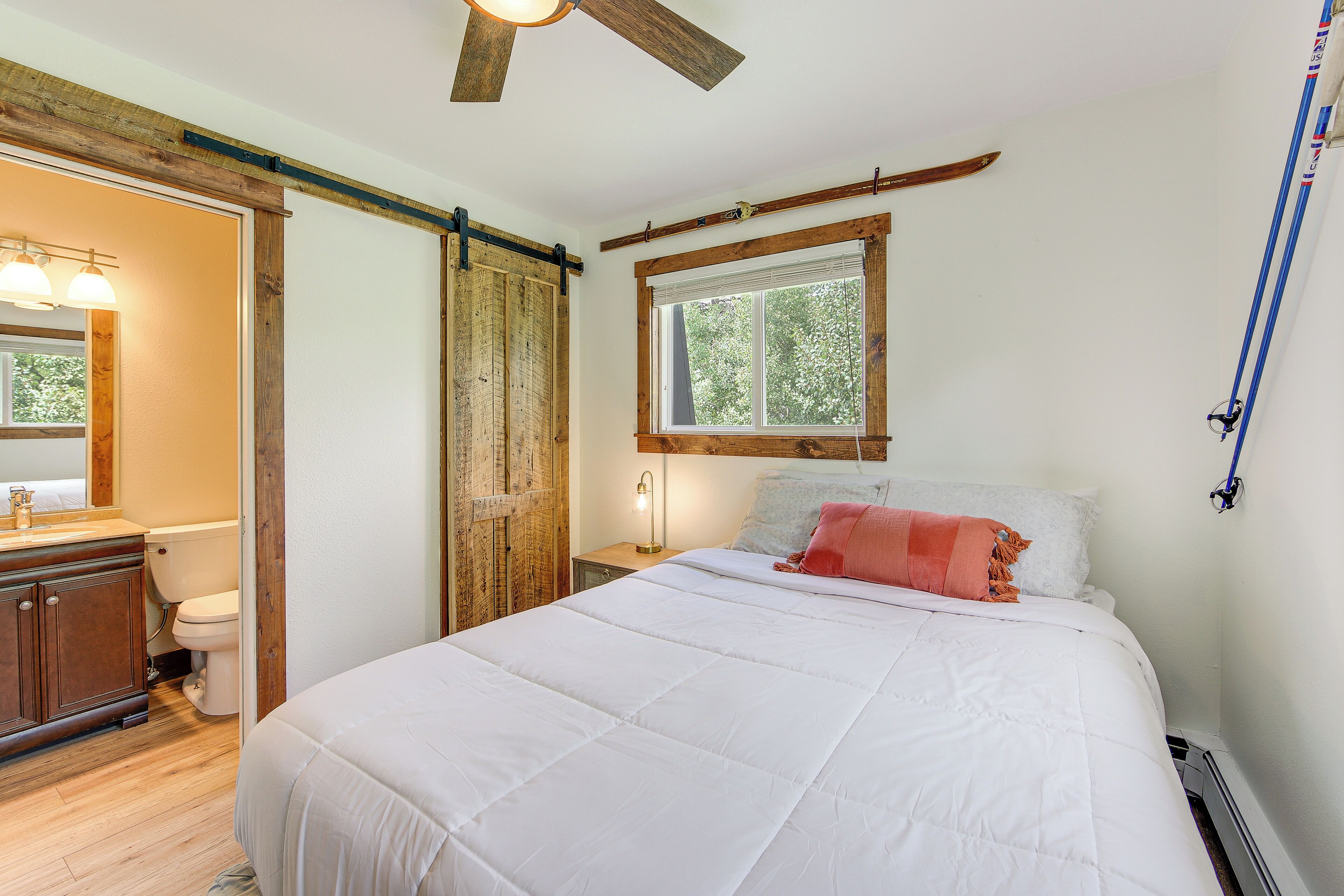 A bedroom with a large bed, a wooden ceiling fan, a window with a view of trees, a bedside table with a lamp, and a wooden wardrobe.