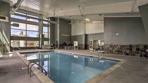 Una piscina interior con paredes de vidrio transparente, suelo de baldosas y una zona de estar con mesas y sillas.