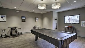 Una mesa de billar en una habitación con un ventanal grande, dos lámparas colgantes y tres taburetes.