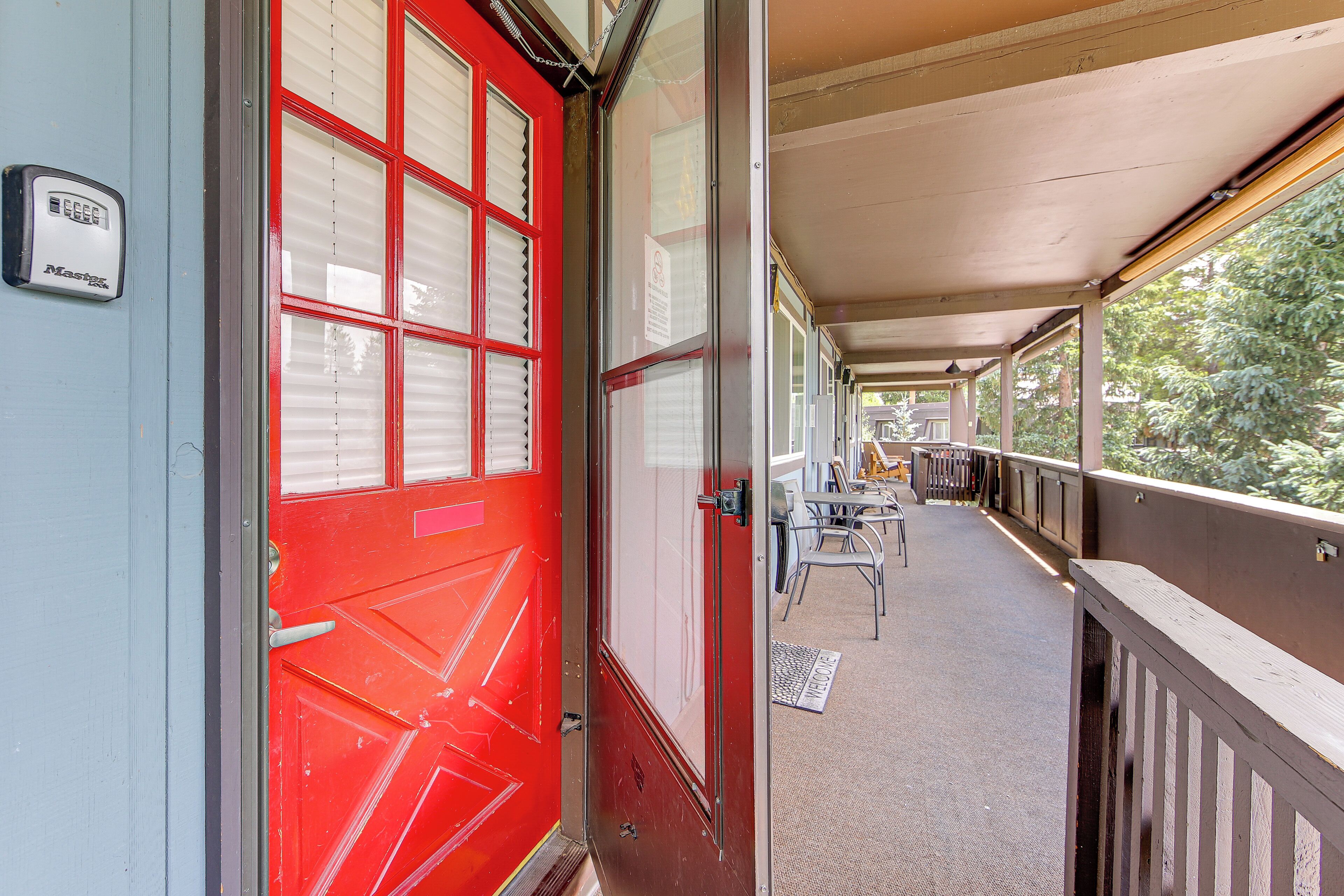 A red door with a security device, a covered outdoor area with tables and chairs, and a view of greenery outside.