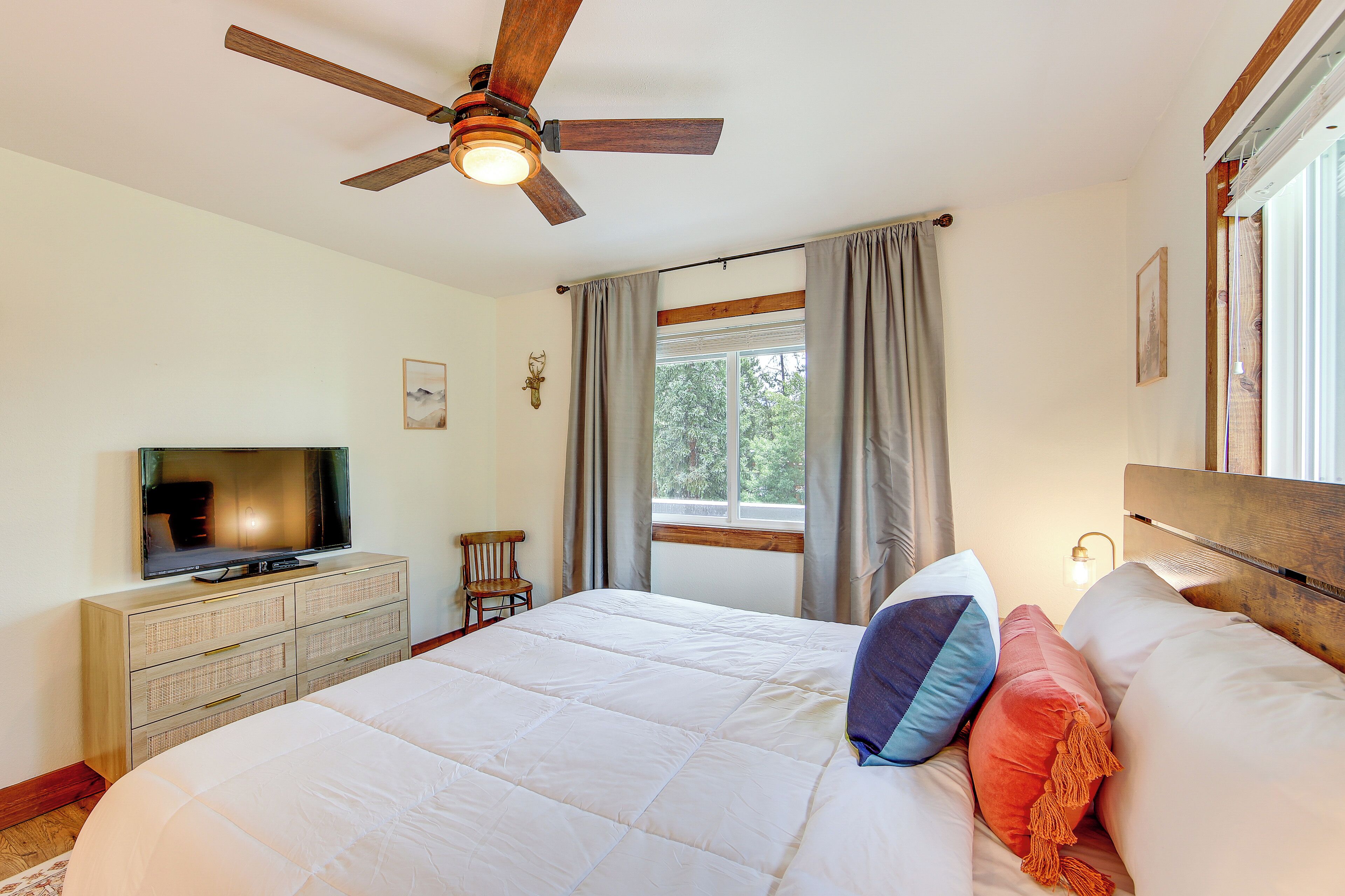 Une chambre à coucher avec un lit, un téléviseur posé sur une commode, un ventilateur de plafond et une fenêtre avec des rideaux.