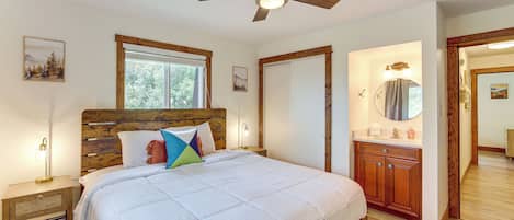 Un dormitorio con una cama grande, cabecera de madera, lámparas de noche y un ventilador de techo, con vistas al exterior a través de una ventana.