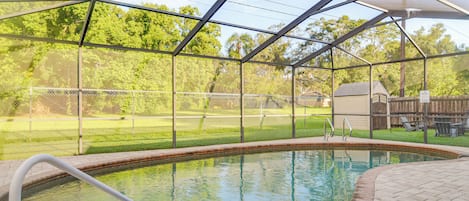 Una piscina techada con techo metálico, rodeada por un patio de ladrillo y una zona verde con una cerca.