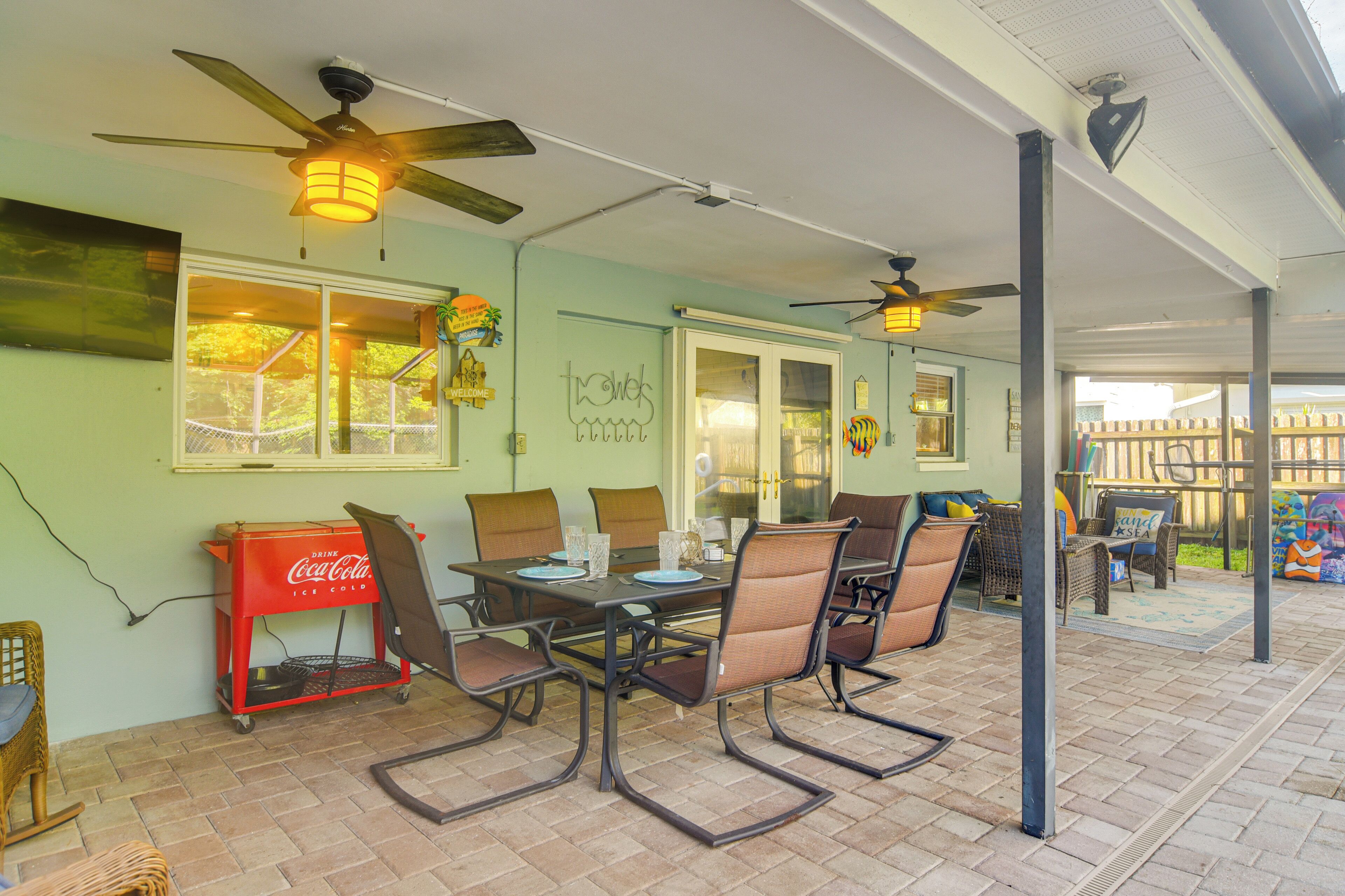 A covered patio area with a dining table, chairs, ceiling fans, and a Coca-Cola cooler.