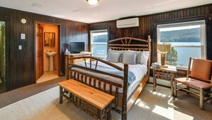Ein Schlafzimmer mit Holzwänden, einem Bett, einer Bank, einem Sessel, einem Fernseher und einem Fenster mit Seeblick.