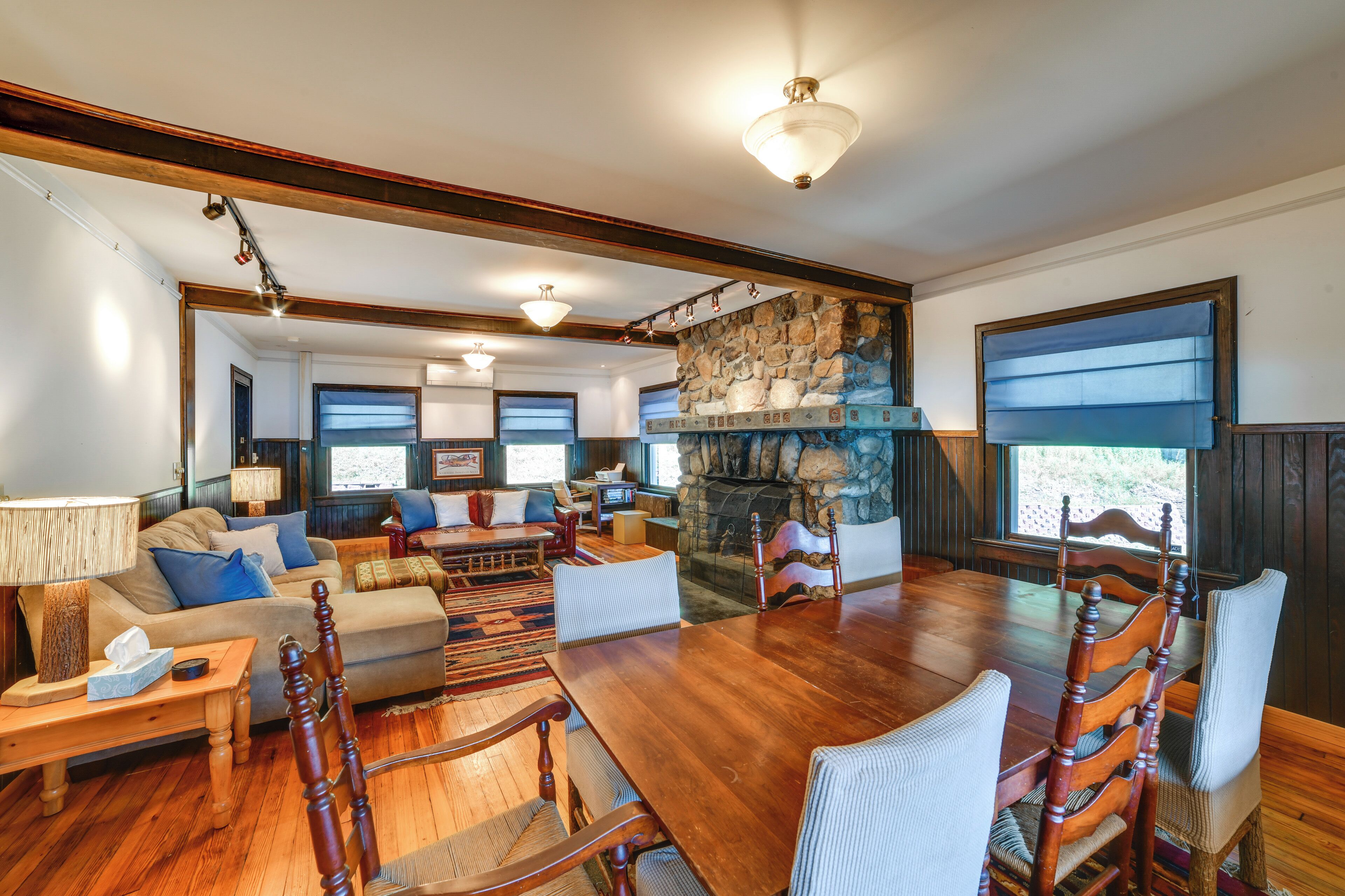 A spacious living room with a large wooden dining table, comfortable seating, a stone fireplace, and large windows.