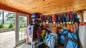 A room with a wooden ceiling and a large window overlooking a brick patio. There are various bags and gear, including a rack of life jackets and a shelf with rolled-up mats. - 'the Nest': Waterfront Lake Ostego Studio (Cooperstown)