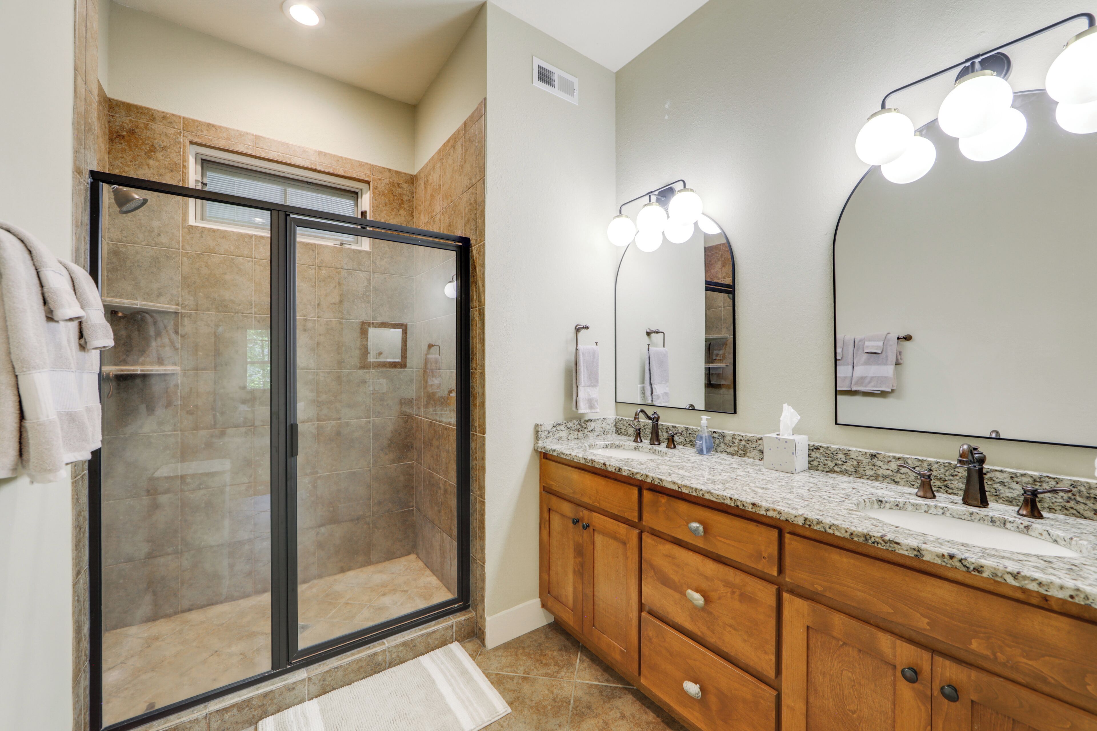 A modern bathroom with a glass-enclosed shower, wooden cabinets, a granite countertop, and a large mirror with lights.