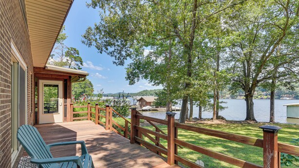 A wooden deck with chairs, a wooden fence, and a view of a lake and buildings in the background. - On Lake Tillery: Spacious Home w/ Screened Porch! (Albemarle)