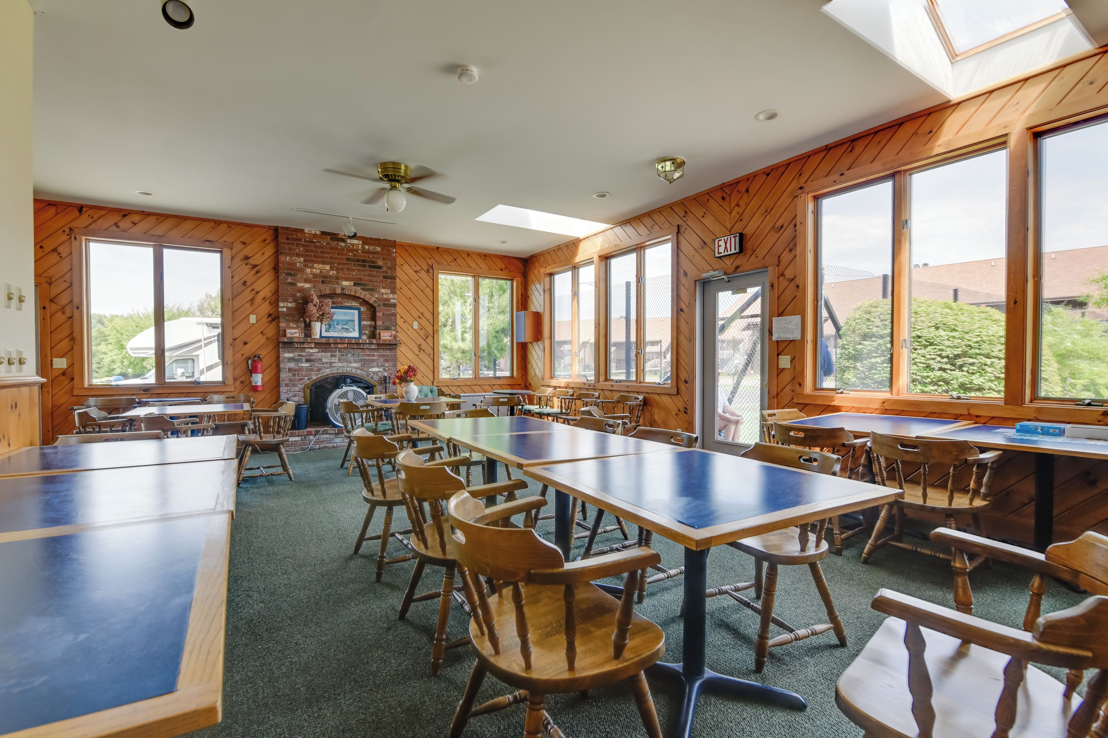 A spacious dining area with long tables and wooden chairs, a brick fireplace, and large windows offering views of the outdoors.