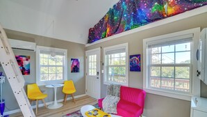A room with a galaxy-themed tapestry, a pink sofa, yellow chairs, and a white coffee table. - 'galactikitty' - Quirky Cabin Near Lake Brownwood! (Brownwood)