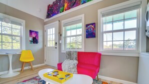 A room with a bright pink sofa, a white coffee table, and a colorful rug. There are two windows with blinds, a ceiling with a galaxy-themed artwork, and a small table with a yellow chair. - 'galactikitty' - Quirky Cabin Near Lake Brownwood! (Brownwood)